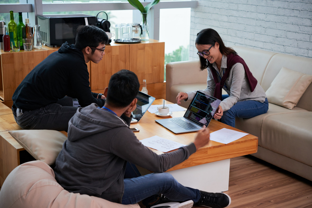 Trio of casual developers collaborate around coffee-table laptops and sketches in a relaxed workspace.