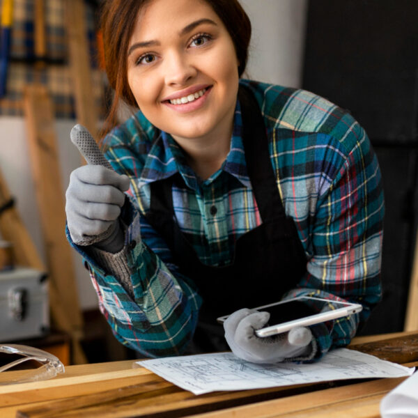Smiling craftswoman in plaid and work gloves gives a thumbs-up over blueprints and a tablet at a woodworking bench.