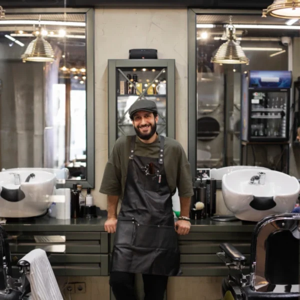 Friendly barber in cap and apron leans against a styling station between two washbasins.