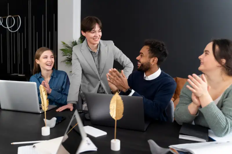 Diverse coworkers applaud and laugh around laptops during an informal office huddle.