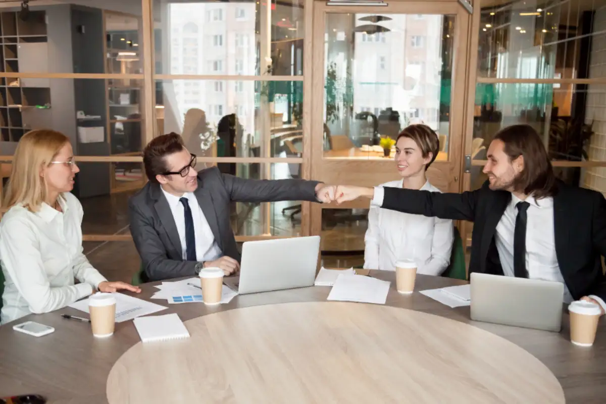 Two suited colleagues fist-bump across a meeting table while teammates look on.