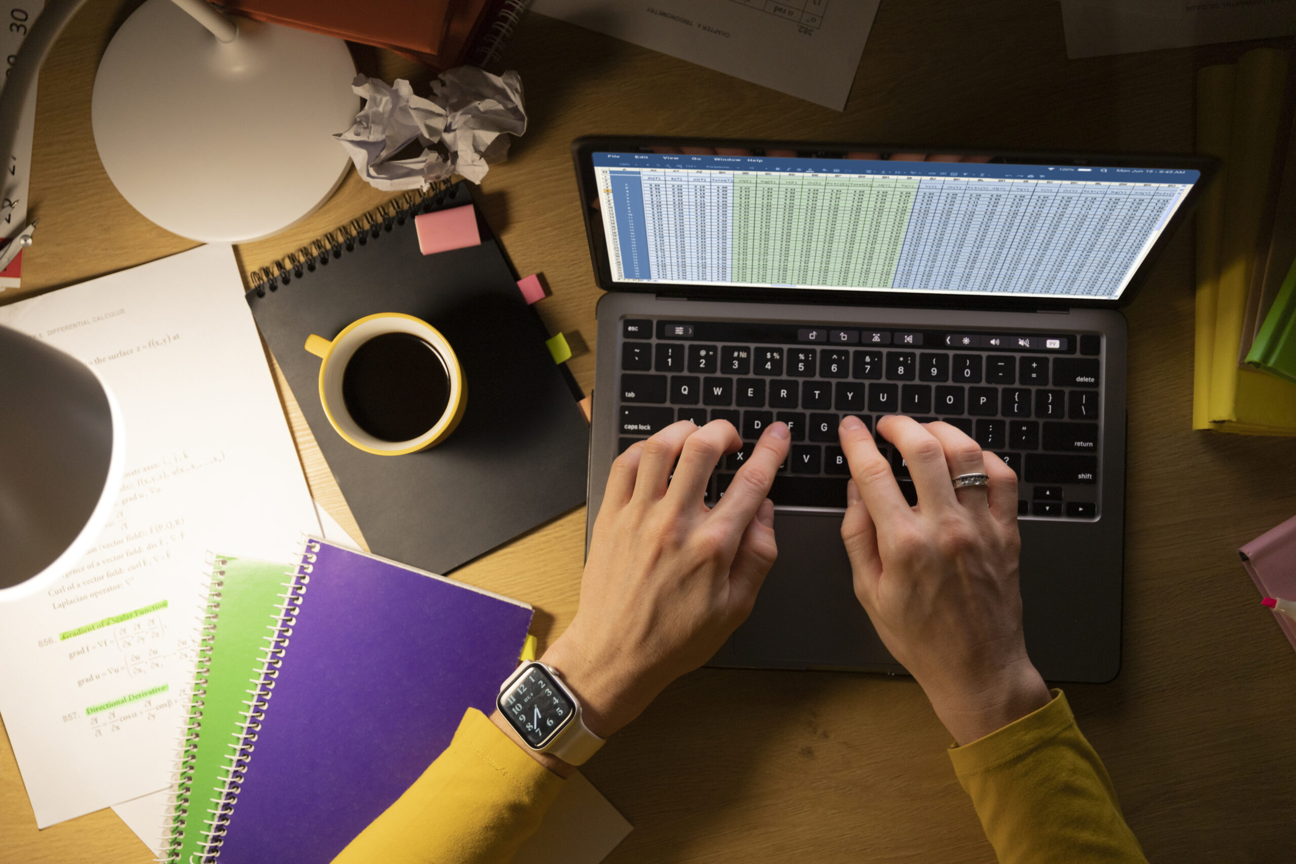 Top-down view of hands typing a spreadsheet on a laptop amid notebooks, papers, and coffee.