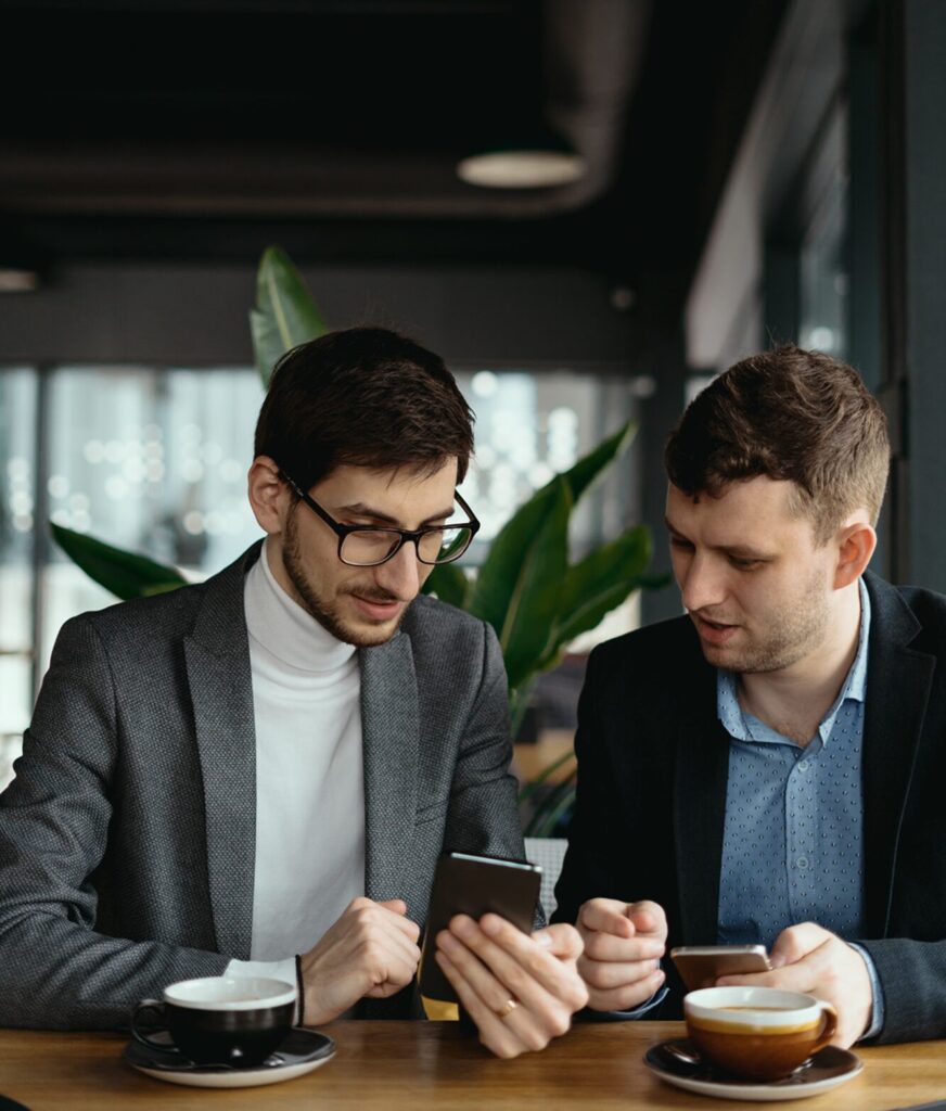 Two business-attired men sit in a café comparing content on their smartphones over coffee.